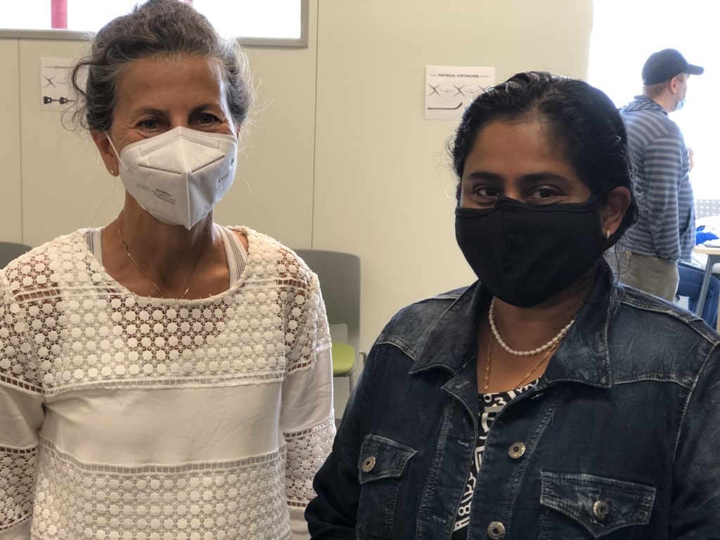 Two women stand smiling with their eyes, as they have face masks on 