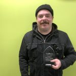 Andrew Baird stands against a green wall holding his award; he wears all black