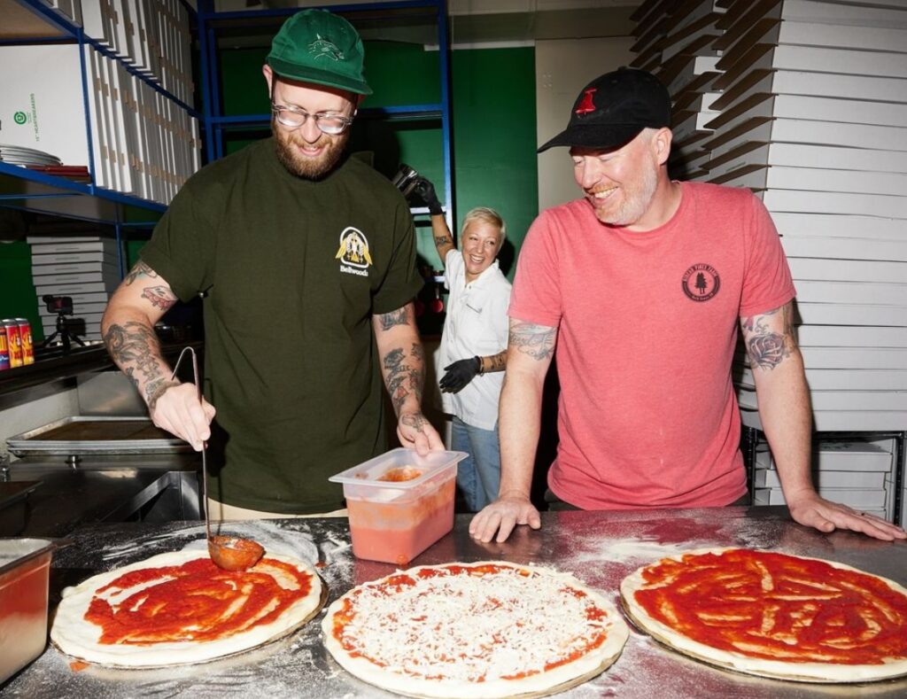 A man in a green t-shirt and hat pours marinara sauce from a ladle onto dough. A woman with short blonde hair smiles in the background, reaching for something.  A man in a red t-shirt watches the other man as he pours on the sauce, smiling. Pizza boxes are stacked in the background.

Un homme vêtu d'un tee-shirt vert et d'un chapeau verse de la sauce marinara à l'aide d'une louche sur de la pâte. Une femme aux cheveux blonds courts sourit à l'arrière-plan, en attrapant quelque chose.  Un homme vêtu d'un t-shirt rouge regarde l'autre homme verser la sauce en souriant. Des boîtes de pizza sont empilées à l'arrière-plan.