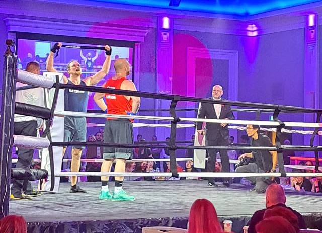 A man holds up a headband in a boxing ring, victorious.